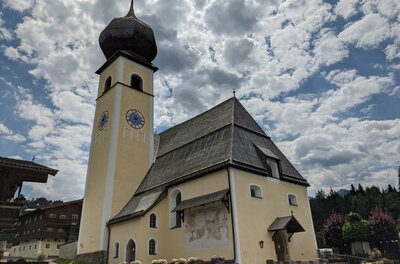 Pfarrkirche in Aurach mit markantem Zwiebelturm und Schindeldach vor bewölktem Himmel.