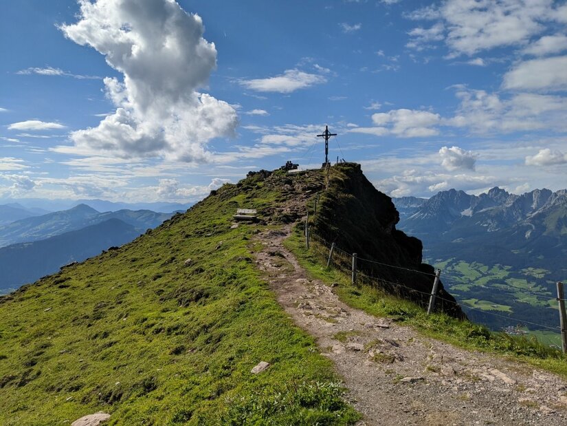 Schmaler Wanderweg führt über einen grasbewachsenen Grat zu einem Gipfelkreuz unter blauem Himmel.
