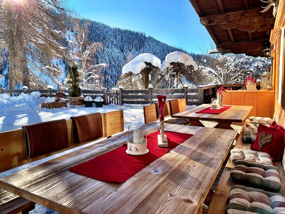 Holztische mit roten Läufern auf einer verschneiten Terrasse vor bewaldeten Bergen.