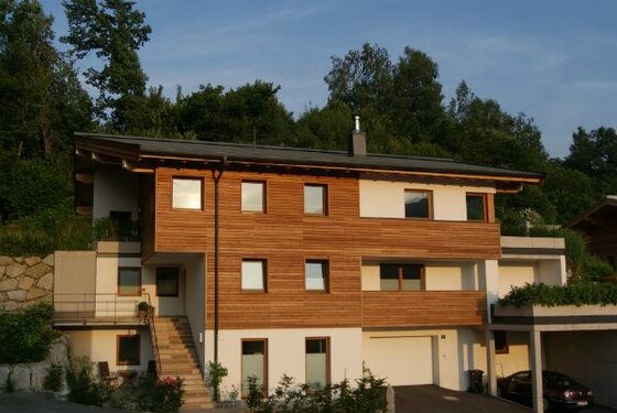Modernes mehrstöckiges Gebäude mit Holzfassade und weißem Putz vor bewaldetem Hintergrund.