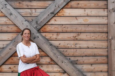 Unsere Kiste Eine Frau in Tracht steht vor einer Holzwand mit dem Schriftzug Unsere Kiste.