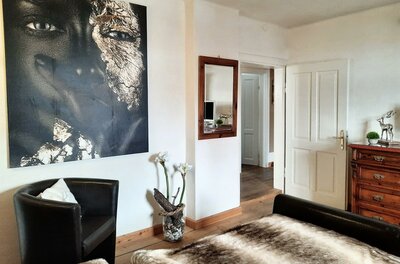 Schlafzimmer mit großem Porträtgemälde, Ledersessel, Holzboden und einer Kommode.