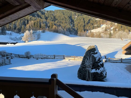 Blick von einem Holzbalkon auf eine verschneite Wiese, einen Zaun und bewaldete Berge im Hintergrund.