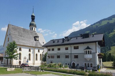 Anton Oppacherplatz Eine malerische Kirche mit einem hohen Turm steht neben einem traditionellen Gebäude. Im Hintergrund sind sanfte Hügel und ein klarer Himmel zu sehen.