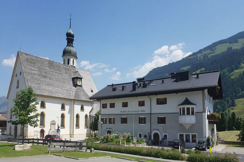 Anton Oppacherplatz Eine malerische Kirche mit einem hohen Turm steht neben einem traditionellen Gebäude. Im Hintergrund sind sanfte Hügel und ein klarer Himmel zu sehen.