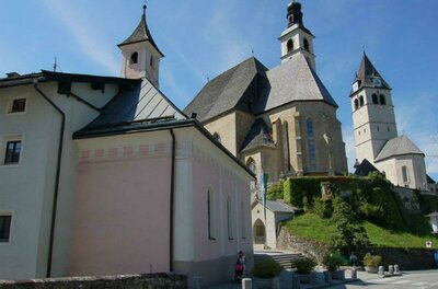 Ein historisches Gebäude mit mehreren Türmen und unterschiedlichen Dachformen. Im Vordergrund ist ein helles Haus zu sehen, das sich an eine grüne Hügelkuppe anschmiegt.