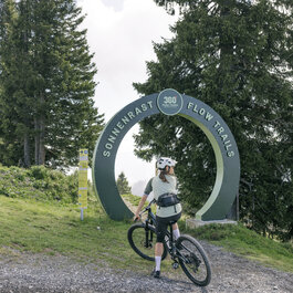 Ein Mountainbiker steht bei den 360 Flow Trails in Kitzbühel vor einem großen Schild, das den Eingang zu den Sonnenrast Flow Trails markiert.