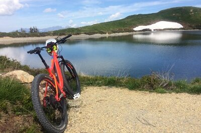 Ein orangefarbenes Mountainbike steht auf einem Schotterweg vor einem Bergsee mit Schneerest am Ufer.