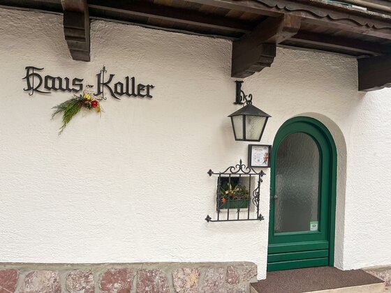 Eingang Pension Haus Koller Weiße Hauswand mit grüner Rundbogentür, Schriftzug Haus Koller, Laterne und kleinem Gitterfenster.