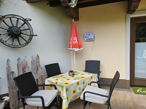 Tamara's Apart Terrasse Außenterrasse mit Tisch, vier Stühlen, Sonnenblumendecke und einem dekorativen Wagenrad an der Wand.