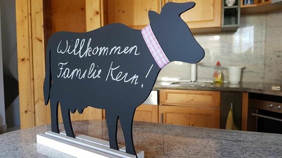 Willkommensschild in Kuhform Schwarze Kreidetafel in Form einer Kuh mit Willkommensgruß auf einer Arbeitsplatte in einer Küche.