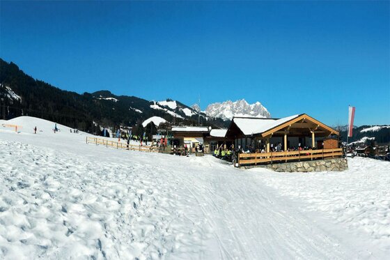 Holzhütte mit Terrasse an einer verschneiten Skipiste vor Bergkulisse.