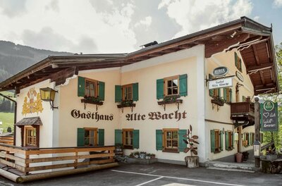 Zweistöckiges Gasthaus Alte Wacht mit hellen Wänden, grünen Fensterläden und Holzbalkon vor bewaldetem Berg.
