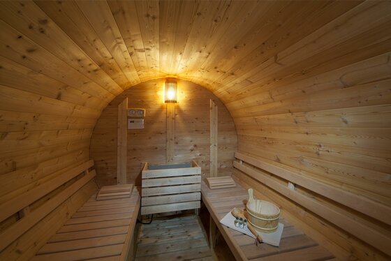 Innenansicht einer runden Fasssauna aus hellem Holz mit zwei Sitzbänken, Saunaofen und Aufgusszubehör.