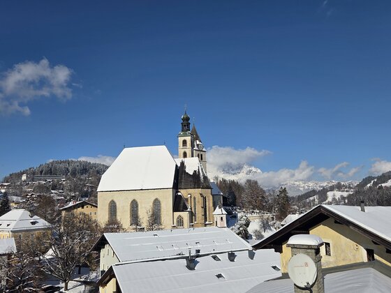 Ausblick Streifblick Lodge Eine schöne winterliche Landschaft mit einer Kirche und schneebedeckten Dächern. Im Hintergrund sind die Berge und ein blauer Himmel zu sehen.