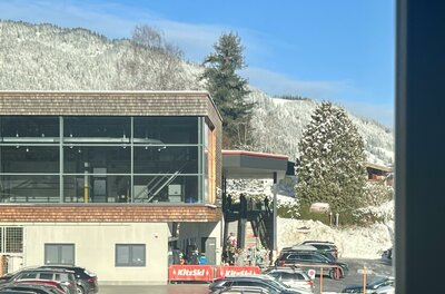 Ein modernes Gebäude mit großen Fenstern, umgeben von schneebedeckten Bergen. Im Vordergrund sind viele parkende Autos zu sehen.