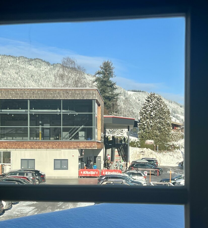 Ein modernes Gebäude mit großen Fenstern, umgeben von schneebedeckten Bergen. Im Vordergrund sind viele parkende Autos zu sehen.