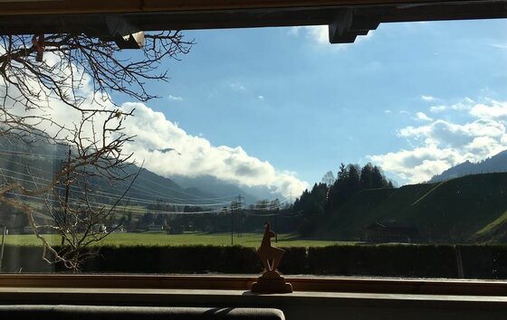 Ein malerischer Ausblick durch ein Fenster auf grüne Wiesen und sanfte Hügel. Im Hintergrund sind Berggipfel und ein blauer Himmel mit einigen Wolken zu sehen.