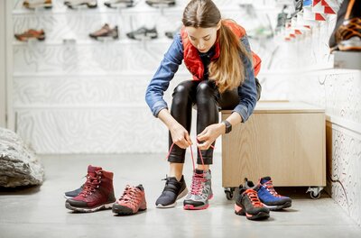 Eine Frau sitzt in einem Sportgeschäft und schnürt einen grauen Wanderschuh mit pinken Akzenten.