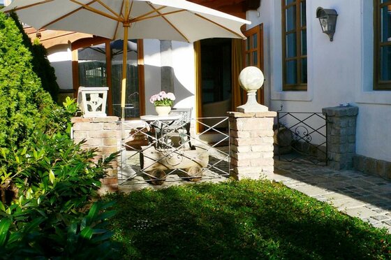 Gepflasterte Terrasse mit weißem Sonnenschirm, kleinem Tisch, Blumen und gemauerten Pfosten vor einem Haus.