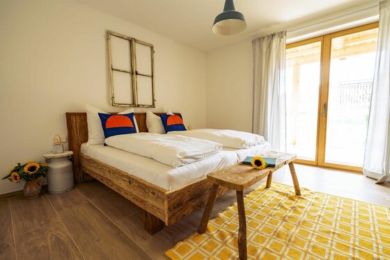 Schlafzimmer mit Doppelbett aus Altholz, gelbem Teppich und Balkonzugang.
