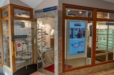 Allmoslechner United Optics Eingangsbereich eines Optikergeschäfts mit Holzrahmen, Glastür und Brillenpräsentationen in beleuchteten Regalen.