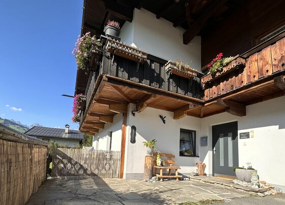 Eingangsbereich Tannenheim Eingangsbereich eines alpinen Hauses mit Holzbalkon, Blumenkästen und einer kleinen Holzbank auf einer Steinterrasse.