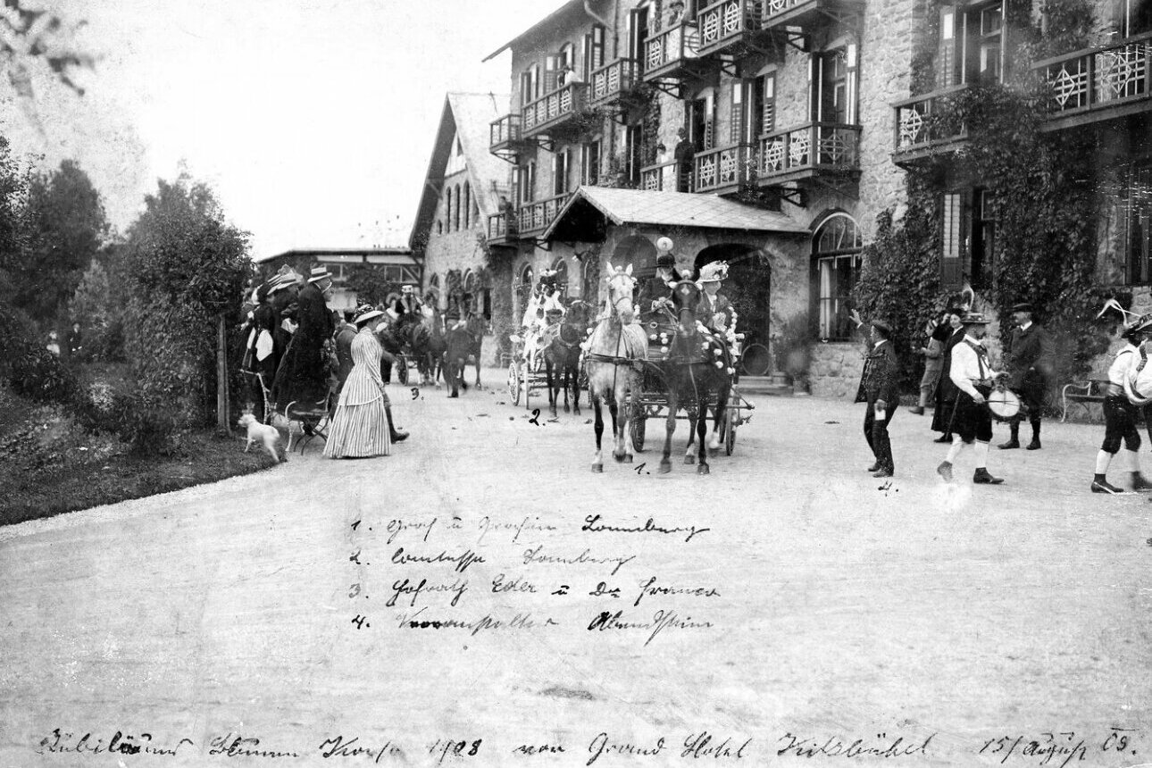 A two-horse carriage and several people are standing in front of the Grand Hotel in Kitzbühel