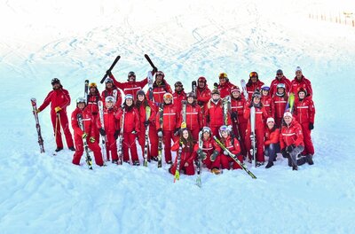 Eine Gruppe von Skilehrern in roten Anzügen posiert mit Skiern auf einer verschneiten Piste.