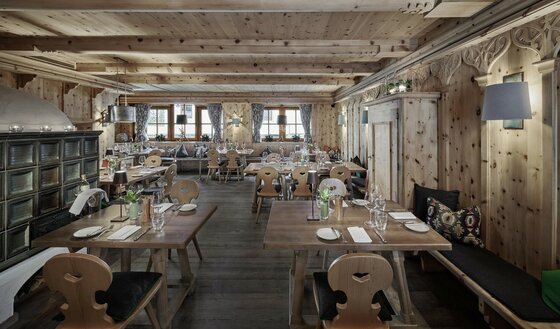 Rustikale Gaststube mit Holztischen, Stühlen mit Herz-Ausschnitt, Kachelofen und holzgetäfelten Wänden.