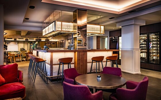 Moderne Hotelbar mit beleuchteter Theke, Barhockern und Sesseln im Innenraum.