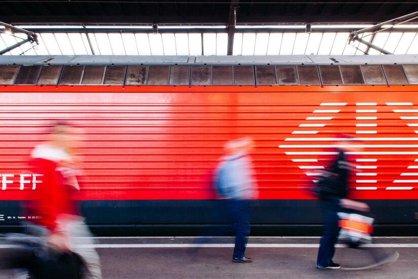 Hahnenkamm Express Ein Bahnhof mit einer roten Wand im Hintergrund. Menschen gehen schnell vorbei, während sie unterwegs sind.