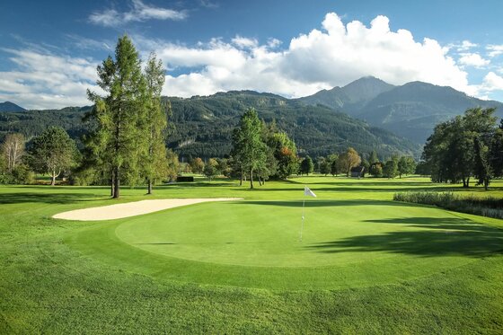 Golfplatz Zell am See Gepflegtes Green auf einem Golfplatz mit Sandbunker vor bewaldeten Bergen und blauem Himmel.