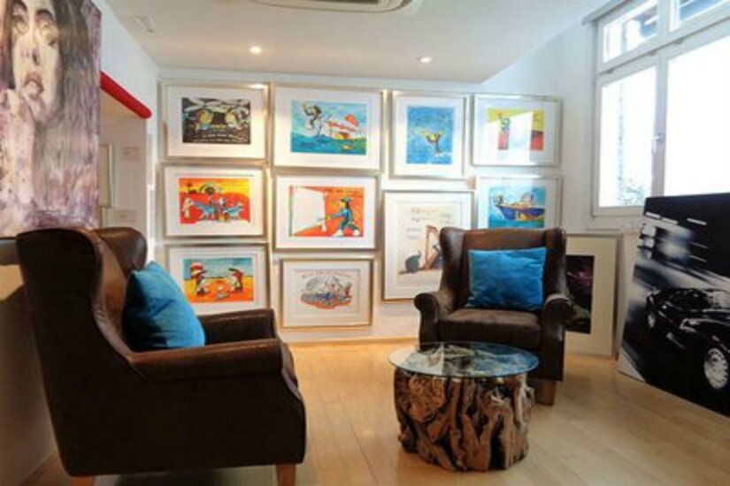 Walentowski Galerie Kitzbühel Eine gemütliche Sitzecke mit zwei Stühlen und einem Glastisch. An der Wand hängen farbenfrohe Kunstwerke in Rahmen.