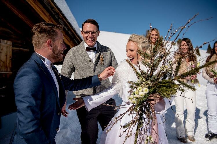 Hochzeitsplaner Finest Wedding Eine fröhliche Hochzeitsfeier im Schnee mit lächelnden Gästen. Die Braut trägt einen schönen Blumenstrauß und wird herzlich begrüßt.
