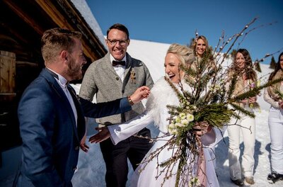 Eine fröhliche Hochzeitsfeier im Schnee mit lächelnden Gästen. Die Braut trägt einen schönen Blumenstrauß und wird herzlich begrüßt.