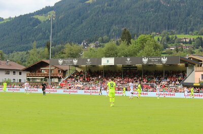 Sportplatz Ein Fußballspiel auf einem Stadionfeld mit Zuschauern im Hintergrund. Die umliegende Landschaft ist grün und bergig.