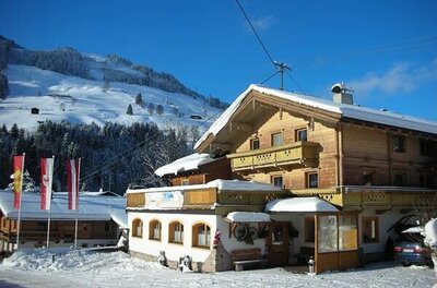 Ein mehrstöckiges Holzhaus mit schneebedecktem Dach vor einem bewaldeten Berghang im Winter.