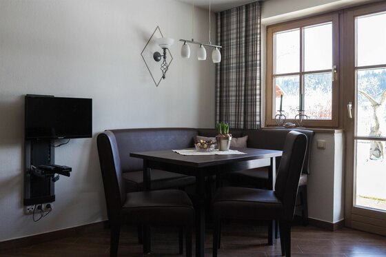 Essecke in der Ferienwohnung Essecke mit dunkler Eckbank, Tisch, zwei Stühlen und einem Wandfernseher neben einem Fenster.