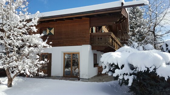 Zweistöckiges Holzhaus mit Balkon und schneebedecktem Dach sowie verschneiten Bäumen im Vordergrund.
