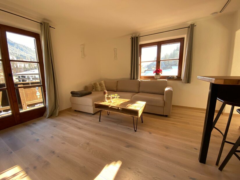 Wohnzimmer im Haus Ingrid Wohnbereich mit beigem Ecksofa, Holztisch und Parkettboden vor einem Fenster.