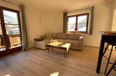 Wohnzimmer im Haus Ingrid Wohnbereich mit beigem Ecksofa, Holztisch und Parkettboden vor einem Fenster.