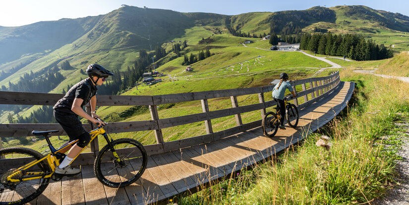 Zwei Mountainbiker fahren bei den 360 Flow Trails in Kitzbühel über eine Holzbrücke, umgeben von grünen Hügeln und Bäumen unter blauem Himmel.