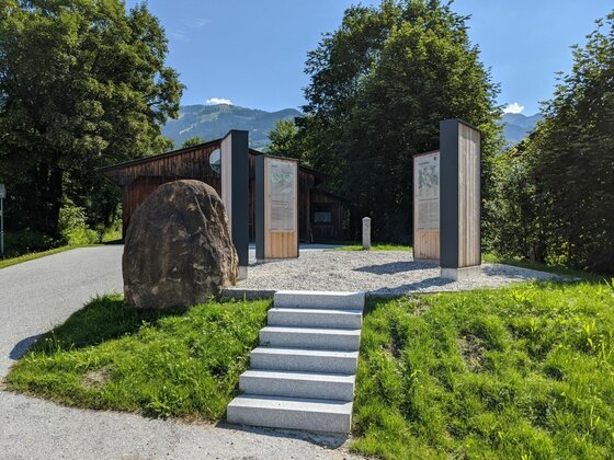 Infoplatz mit Steinstufen, Informationstafeln aus Holz und Metall sowie einem großen Findling vor Bäumen.