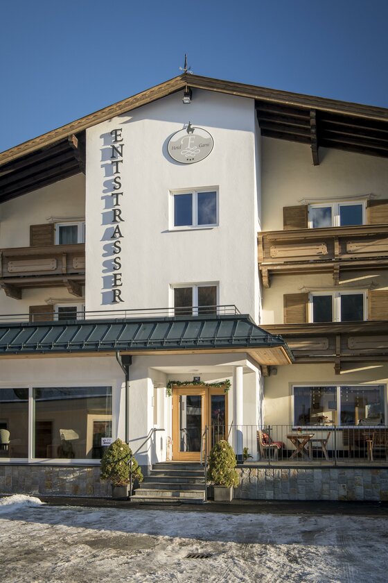 Aussenansicht Winter 2 Vorderansicht des Hotel Garni Entstrasser mit Holzbalkonen und Eingangsbereich im Winter.