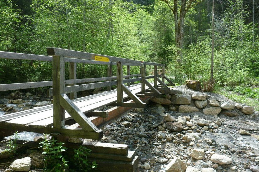 Eine Holzkonstruktion über einen kleinen Bach, umgeben von grünen Bäumen. Der Weg lädt zu einem ruhigen Spaziergang in der Natur ein.