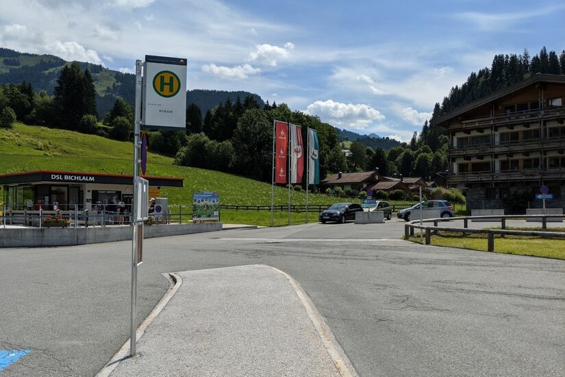 Bushaltestellenschild vor einem Parkplatz mit Bergen im Hintergrund.
