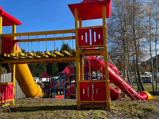 Spielplatz am Campingplatz Bunter Spielplatz mit Klettertürmen, Hängebrücke und Rutschen vor Bäumen unter blauem Himmel.