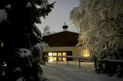 Lutzenberg 19 im Winter Beleuchtetes Gebäude im Schnee bei Dämmerung mit verschneiten Tannen im Vordergrund.