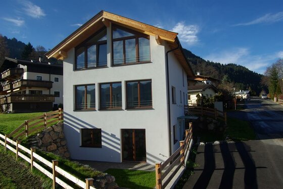 Modernes dreistöckiges Wohnhaus mit weißer Fassade, Satteldach und großen Fensterfronten vor bewaldeten Bergen.